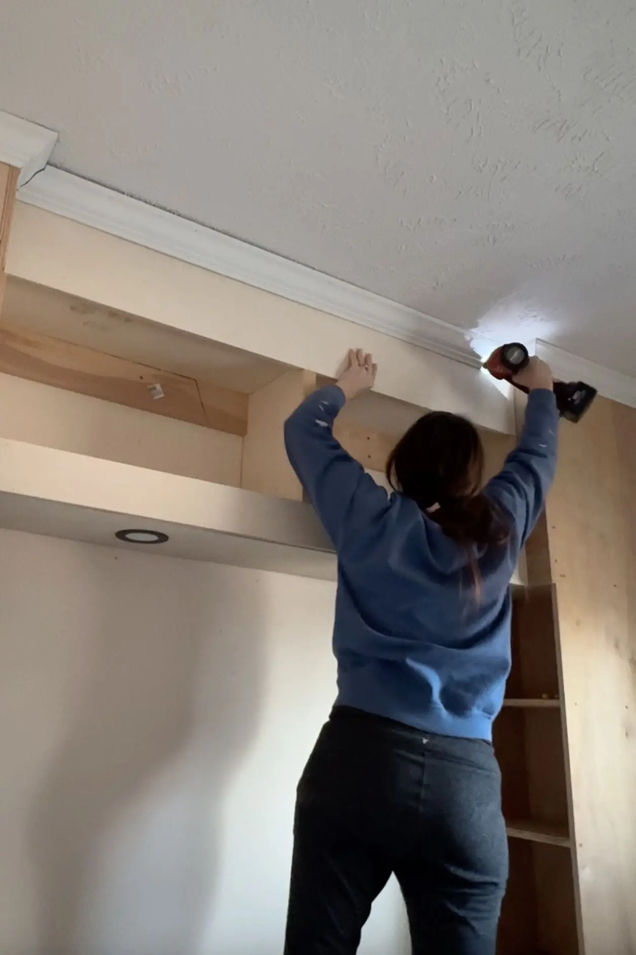 A DIYer attaching white crown molding to the top of a built-in bedroom unit using a brad nailer. She is reaching up to secure the trim along the ceiling, adding a polished detail to the project.